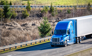 Blue bright big rig semi truck transporting goods in dry van semi trailer driving on the interstate...