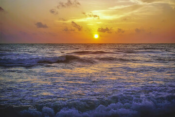 Sunset over the Indian Ocean. The sky is colored orange. Light clouds. Surf waves roll onto the beach with foam and spray. Shri Lanka