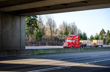 Red popular big rig semi truck transporting lumber wood on step down semi trailer running under the...