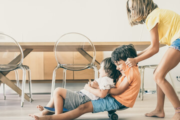 Cheerful children playing with skateboard at home. Blonde adorable girl pushing her two playful...