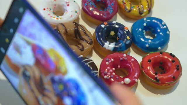 A close-up video is taken on a smartphone. On a white table lie donuts covered with glaze of different colors and sprinkled with colored sprinkles. Photo concept for social networks.