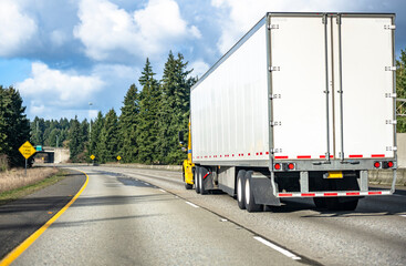 Yellow bonnet big rig semi truck transporting cargo in dry van semi trailer running on the turning wide highway road with evergreen trees on the side and bridge ahead