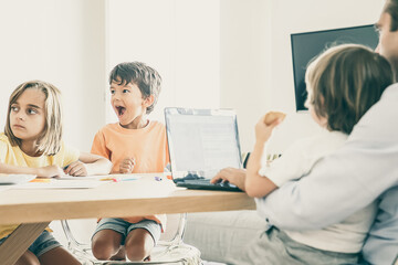 Happy kids drawing doodles when dad working on laptop and holding son on knees. Exited children painting on paper. Caucasian family sitting at table. Childhood, creativity and weekend concept