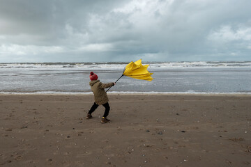 Young boy struggling with large yellow umbrella on beach in stormy weather. Winter beach scene.