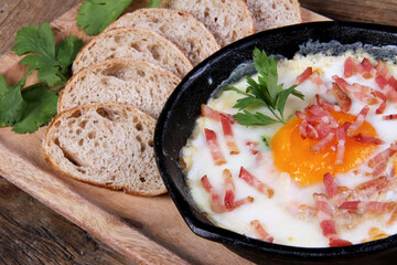 egg with bacon in black casserole with sliced bread on wooden board