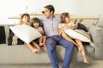 Caucasian dad sitting on sofa and embracing cute kids. Loving middle-aged father relaxing with adorable children on coach in living room and talking. Childhood, family time and fatherhood concept