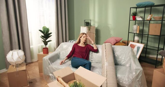 Pretty Caucasian tired young woman moving into new apartment unpacking stuff falling on couch after relocation. Female resting on moving day in room with many carton boxes parcels