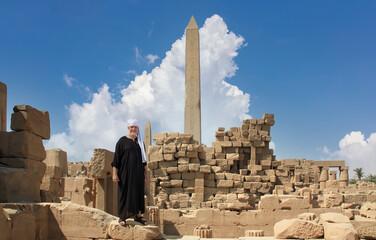 An elderly Egyptian in a black robe and a turban stands in the area of the temple complex in Karnak...