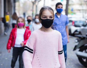 Confident tween girl in black face mask walking along city street in autumn day. Concept of Covid...