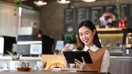 Smiling young female freelance working on digital tablet and sitting in modern coffee shop.