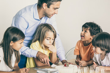 Caucasian father cutting delicious cake for children. Cute little kids surrounding table, celebrating birthday together, talking and waiting for dessert. Childhood, celebration and holiday concept