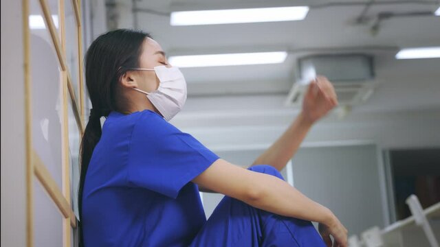 Young Asian woman nurse wearing uniform and surgical mask looking distraught and sad sitting on the floor while taking break after hard work in hospital - 4k 60fps footage