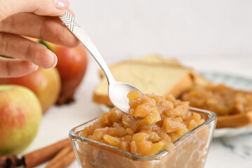 Female hand with spoon and bowl of sweet apple jam on light background