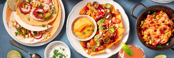 Mexican food panoramic header on a blue background. Nachos with chili con carne, tacos with chicken, and various dips, top shot