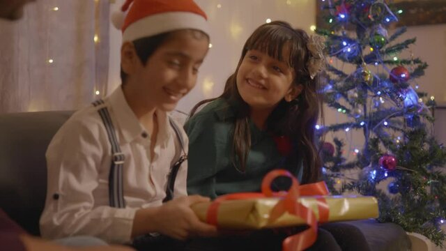 shot of cheerful indian children receiving present wrapped up in gift box from their father or parent on the occasion of christmas eve