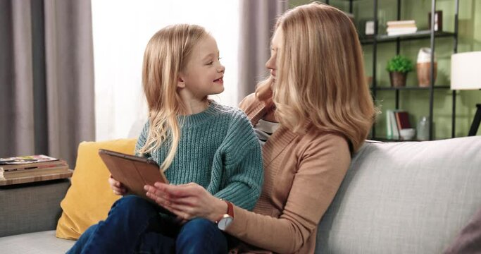 Portrait of cheerful smiling Caucasian caring mom spending time with little cute daughter teaching her typing on tablet device, browsing online sitting on sofa. Motherhood concept. Happy childhood