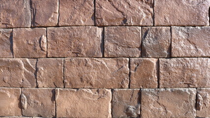 surface lined with rough natural stone of red-brown color with preserved relief texture, stone backdrop of the fragment's exterior in sunlight, emphasizing the details of cracks and bulges