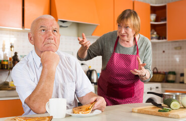 Upset and tired senior man sitting at home table during lunch on background with disgruntled wife scolding him