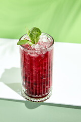 Black currant lemonade drink over green background. White table with sunlight and palm leaf hard shadow. Summer, tropical and juicy concept