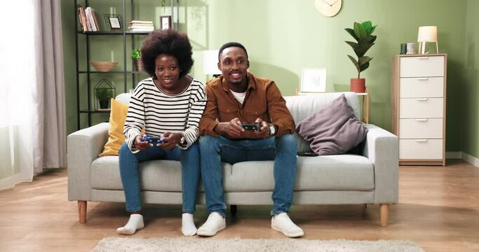 African American happy young married couple playing video games sitting on sofa at home. Handsome man loses to his pretty wife the game on console. Gamers having fun. Rest concept