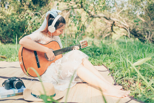 Girl wearing headphone to listen music and playing guitar in the forest. Music hobby and picnic concept.