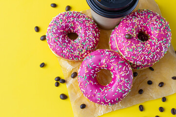 Yummy pink glaze donuts with colorful sprinkles, takeaway coffee cup and roasted coffee beans on yellow background. Junk food.