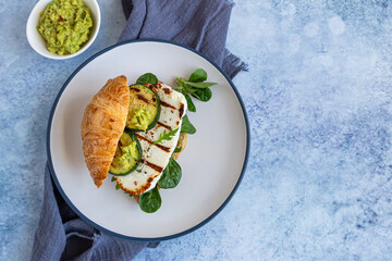 Croissant sandwich with grilled halloumi cheese, arugula and grilled zucchini, blue concrete background. Healthy breakfast or lunch.