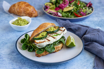 Croissant sandwich with grilled halloumi cheese, arugula and grilled zucchini, blue concrete background. Healthy breakfast or lunch.