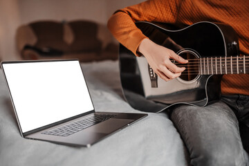 Man learning to play guitar with the help of online learning at home. Guy sitting on bed with...