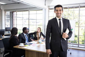 Manager in a meeting room with subordinates sitting in the background. A young man in a black suit stands confident in his leadership character.