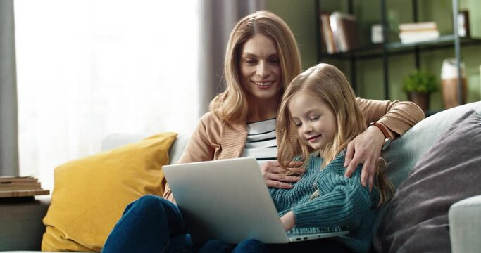 Happy mom with little daughter sitting hugging on sofa at home and watching cartoons online using laptop.