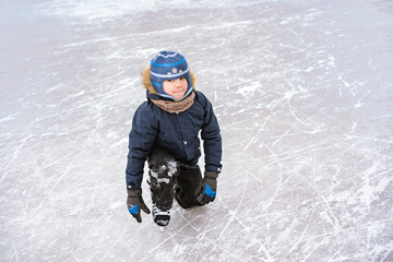 Fototapeta na wymiar cute caucasian preschooler boy learn to skate on ice rink. Kid wearing winter jacket, hood hat and neckwarmer standing on one knee. Image with selective focus