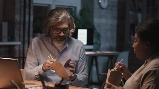 Medium footage of multi-ethnic couple of office workers staying late in office, talking and eating noodles together