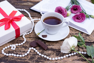 Fototapeta na wymiar Morning time, beautiful picture for blog post in Instagram. Relax with cup of coffee with book and chocolate, decorate by flowers and beads, white gift box. Enjoy your free minutes. Wooden background