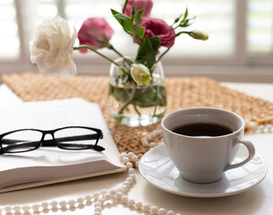 Beautiful composition for blog post in Instagram. Cup of coffee, open book, flowers in low glass vase and beads on table with brown napkin. Free minute at work and pleasure from rest. Close-up