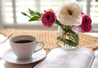 Beautiful composition for blog post in Instagram. Cup of coffee, open book, flowers in low glass vase and beads on table with brown napkin. Free minute at work and pleasure from rest. Close-up