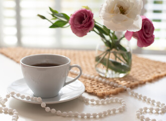 Fototapeta na wymiar Unusual, beautiful composition for posting on Instagram. Cup of coffee, flowers in low glass vase and beads on brown napkin background on table. Free minute at work and pleasure from rest. Close-up
