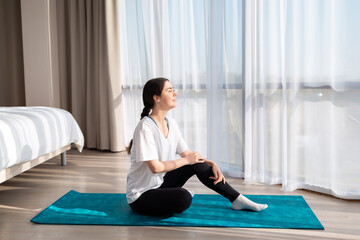 Yoga. A young beautiful woman is sitting on a sports mat, looking out the windows and smiling at the morning sun. Side view. Copy space. The concept of home workouts