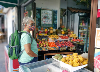 Senior woman with gray hair and a protective mask against the coronavirus stops in a shop and looks at the tropical fruits displayed in the boxes