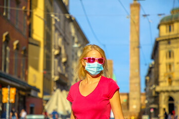 Fashion woman with a surgical mask and sunglasses with Two Towers building at sunset in Bologna town. COVID-19 Coronavirus after quarantine lockdown of Bologna city in Italy.