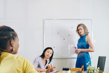 Beautiful serious young businesswoman standing at whiteboard and listening to question of colleague after drawing chart