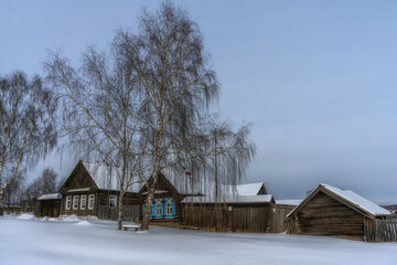 A rural street in winter with old wooden houses, a slanting plank fence and a bench between two tall birches in the old Ural village of Shurala (Russia). a lot of pure white snow, cloudy sky 