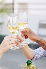 Close-up image of women clinking glasses of white wine over bottle wrapped in tinsel when...