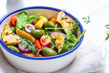 Green salad with artichokes, tomatoes and olives in white bowl.