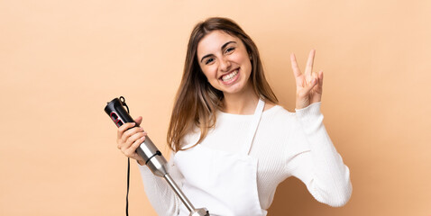 Fototapeta na wymiar Woman using hand blender over isolated background smiling and showing victory sign