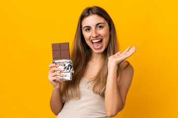 Young woman over isolated yellow background taking a chocolate tablet and surprised