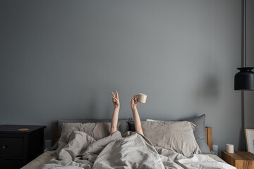 Woman laying in bed and holding mug with coffee with hand and show "Victory" symbol. Happy morning concept. Modern interior design with bedroom, bed, crumpled linen, pillows against grey wall.