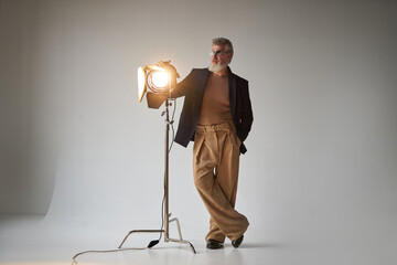 Full length shot of classy middle aged man in glasses looking aside, standing next to studio spotlight while posing for camera over white background
