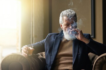 Portrait of elegant mature businessman smoking cigar and drinking whisky while relaxing in a chair...