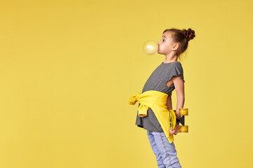 happy little child girl with yellow skateboard blowing a big bubble gum over yellow background....